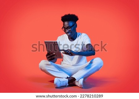 Happy young african american guy in eyeglasses sitting on floor, using modern pc digital pad, male digital nomad watching content online, using great mobile app on red in neon light, copy space