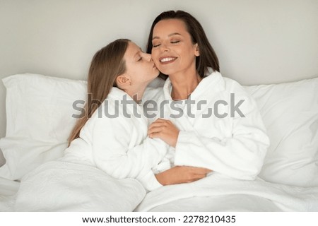 Smiling little caucasian girl kiss on cheek of young woman in bathrobe, relaxing on white soft bed, enjoy spare time, beauty care in bedroom interior. Love, relationship mom and daughter at home