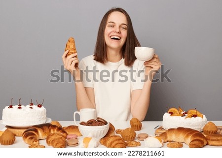 Indoor shot of smiling delighted woman with brown hair sitting at table isolated over gray background, drinking tea with croissant, enjoying her dinner, laughing happily.