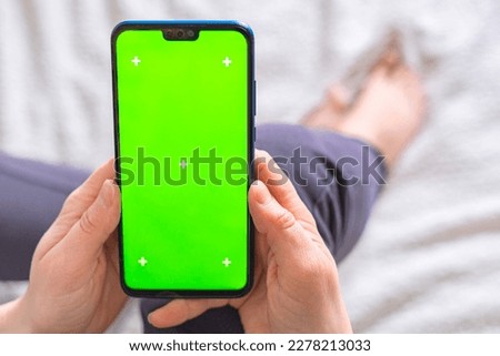 Woman hands hold a smartphone with green screen and sits on the bed background. Chroma key phone.