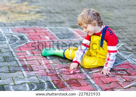 Creative leisure for kids: adorable kid boy clothing as firefighter having fun with fire truck picture drawing with chalk, outdoors. Dreaming of future profession.