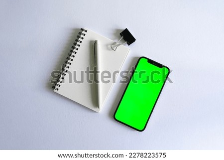 Smartphone mock up with green screen beside a notebook and a pen on a white background desk