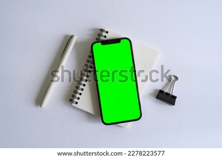 Smartphone mock up with green screen beside a notebook, a pen and a black clip on a white background desk