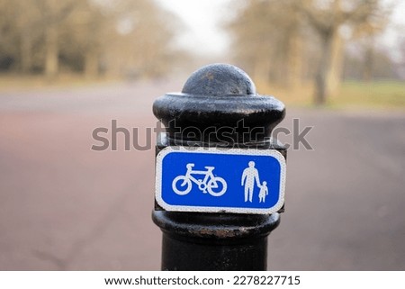 Black painted bollard in public park in street road with blue and white sign with bicycle and person holding a childs hand symbol to warn pedestrians and cyclists of traffic on paved walkway