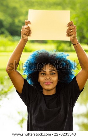 GOIANIA GOIAS BRAZIL - MARCH 20 2023: A young woman, with dyed blue hair, holding a blank sign, with a landscape in the background.