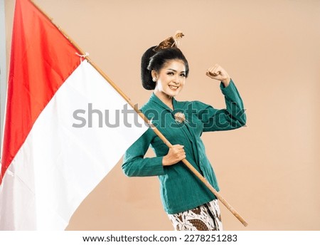 asian woman in green kebaya standing holding the indonesian flag and clenching her arm on isolated background
