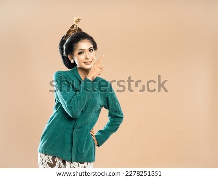 smiling asian woman in green kebaya looking and pointing on her left side while put her hand on her waist on isolated bacground
