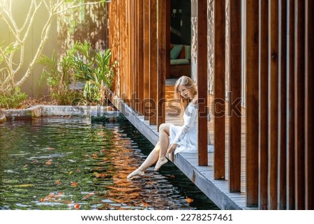 Woman relax in luxury spa with fish pond, enjoy bright morning and nature, relaxing and having good time sitting on wooden bridge barefoot. female playing feet in water at outdoor pond. Slow motion