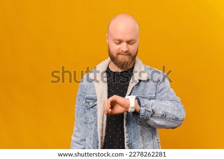 Cool smiling bearded bald man wearing jeans jacket is checking hi watch over yellow background.