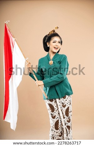 asian woman in green kebaya standing holding the indonesian flag with her both hands on isolated background