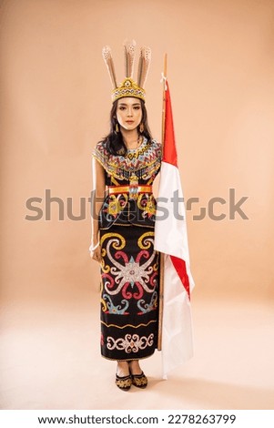 asian woman in one set of traditional clothes of dayak tribe standing forward with solemn face and holding the indonesian flag on isolated background