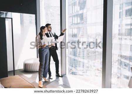 Side view of young office coworkers standing near glass wall in modern office and looking away at high rise buildings and discussing with documents in hand Royalty-Free Stock Photo #2278263927