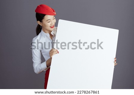 smiling stylish asian female flight attendant in red skirt and hat uniform with blank billboard isolated on gray background.