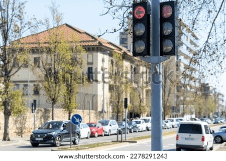 A bustling city street, lined with trees and a variety of architectural structures. A motor vehicle passes under a traffic light while pedestrians go about their day.