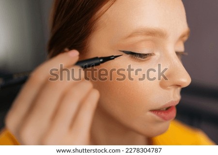 Closeup face of attractive young woman using contour brush drawing arrows on eyelids looking at reflection in mirror. Beautiful redhead female applying eyeliner drawing cat eyes makeup sitting on bed. Royalty-Free Stock Photo #2278304787