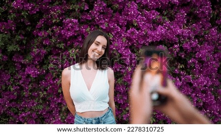 Beautiful woman with freckles and dark loose hair wearing white top poses wall in bloom with purple flowers. Pretty girl is photographed on smartphone