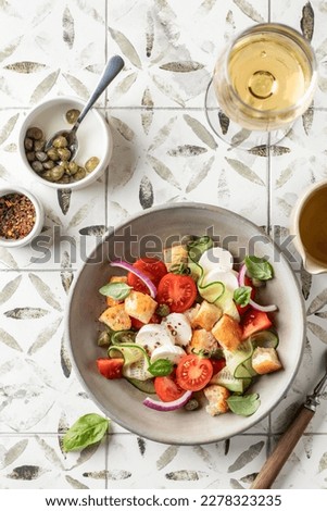 Traditional italian tomato salad panzanella with mozzarella, capers, red onion, croutons, cucumbers and basil with fork and wine glass. Summer salad on printed tile background top view. Vertical