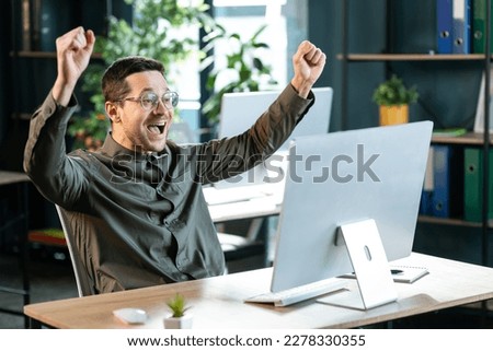 Young guy making YES gesture in front of computer at office. Millennial man reaching success, working with portable pc, making profitable deal, signing online contract, celebrating achievement