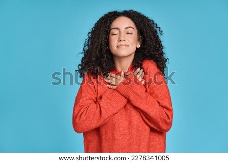 Young adult smiling latin woman, happy pretty curly female model holding hands on chest expressing gratitude, warmth, kindness, peace on mind feeling thankful and touched isolated on blue background. Royalty-Free Stock Photo #2278341005