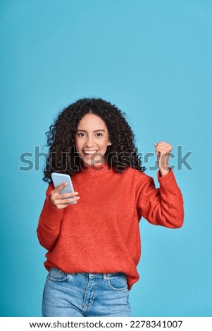 Young happy latin woman winner holding mobile cell phone isolated on blue background. Excited female customer using smartphone apps winning online celebrating discount gift voucher with yes gesture. Royalty-Free Stock Photo #2278341007