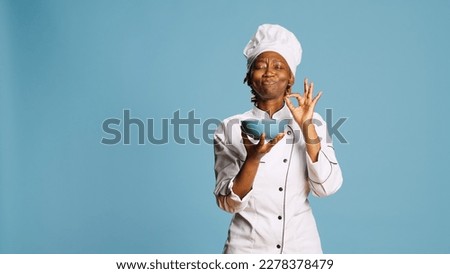 Cheerful gourmet chef smelling food aroma from bowl on camera, enjoying professional meal cooked. Professional female cook savoring tasty food, flavor and scent in studio. Catering service.