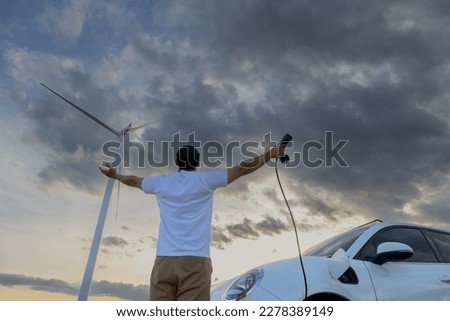 Progressive man spread arm with electric car recharge energy from charging station on green field powered by wind turbine concept of future sustainable energy.