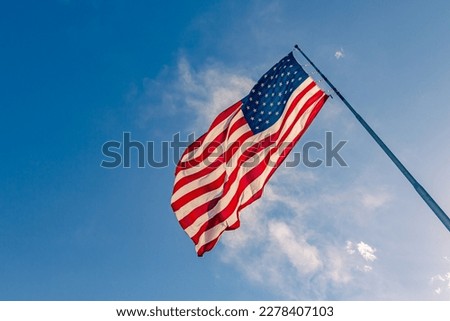 American USA flag on a flagpole waving in the wind. USA Flag Waving United States of America Flag Flying. American flag flying high on a pole against blue sky background on a clear day Royalty-Free Stock Photo #2278407103
