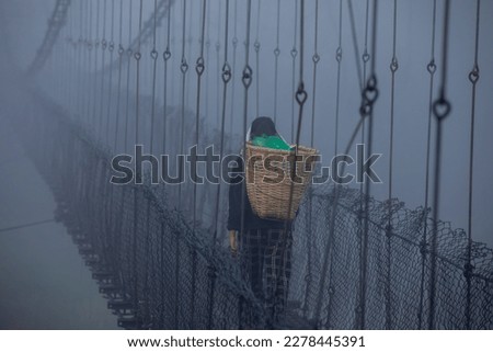 Early dawn, a chilly, foggy winter day a Nepalese woman walking along the suspension bridge carrying a bamboo basket.