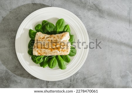Cod fillet baked with garlic butter sauce served with fresh spinach close-up on a white plate, flat lay with copy space