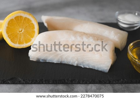 Fresh raw cod fillet, seasoning, olive oil, and lemon slices close-up on a black serving board on the kitchen table