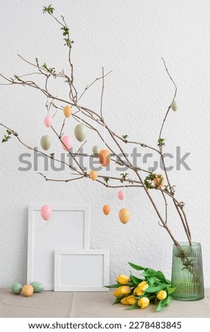Vase with tree branches, Easter eggs, tulip flowers and blank picture frames on table near light wall