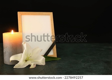 Blank funeral frame, burning candle and lily flower on dark table