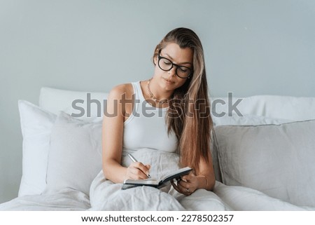 Young brunette caucasian woman in white t-shirt sits on bed writes in diary planning day, agenda. American student girl in glasses makes note, awakes home at bedroom. Daily routine, women. Royalty-Free Stock Photo #2278502357