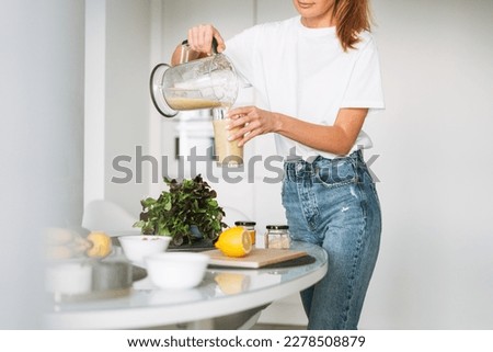Young slim woman in white t-shirt and blue jeans pouring fruit smoothie healthy food in kitchen at home