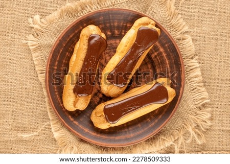 Three chocolate eclairs with a clay plate on a jute cloth, close-up, top view.	 Royalty-Free Stock Photo #2278509333