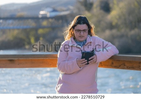 photo of a beautiful middle-aged lady in a pink sweater and light blue pants looking at the phone on the banks of the river at sunset