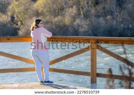 photo of a beautiful middle-aged lady in a pink sweater and light blue pants pensively on the banks of the river at sunset