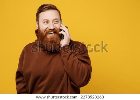 Young redhead caucasian man wearing brown hoody casual clothes talk speak on mobile cell phone conducting pleasant conversation isolated on plain yellow background studio portrait. Lifestyle concept