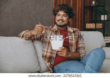 Young Indian man wears casual clothes eat Chinese food cuisine and takeaway carton container box sits on grey sofa couch stay at home hotel flat rest relax spend free spare time in living room indoor