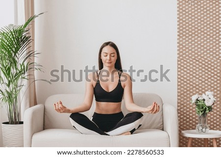 A young woman sits in a lotus position and meditates. Yoga classes at home on the couch. Breathing practices to relieve stress and anxiety Royalty-Free Stock Photo #2278560533