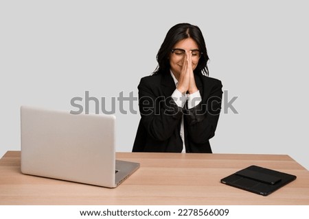 Young indian woman in a table with a laptop and tablet isolated holding hands in pray near mouth, feels confident.