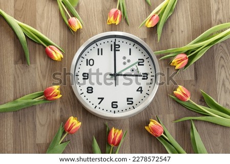 A clock shows three hours. Tulips are lying around. A symbol of the change of time. Daylight saving time. Moving the hands forward. The green arrow indicates the direction of movement of the hands. Royalty-Free Stock Photo #2278572547