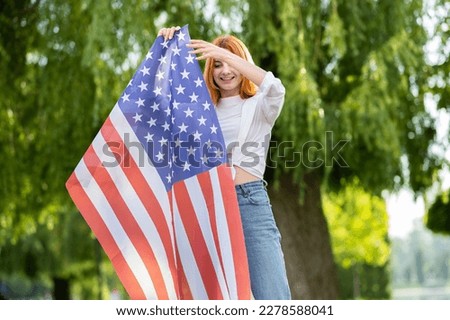 Happy teenage girl posing with USA national flag standing outdoors in summer park. Positive young woman with United States banner outdoors.
