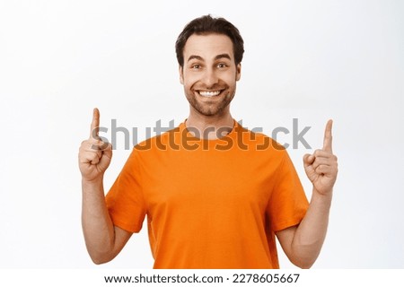 Portrait of man with beard and white smile, points fingers up, shows promo, logo upwards, stands in orange t-shirt. Royalty-Free Stock Photo #2278605667