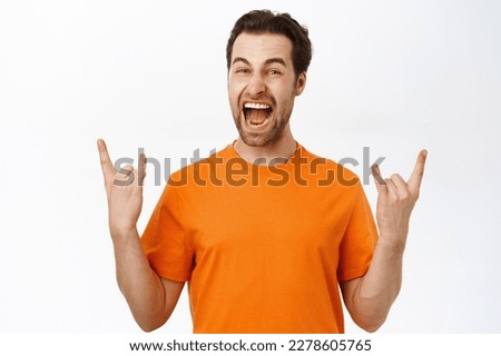 Handsome happy guy shows rock on, heavy metal sign, enjoying smth, standing over white background in orange tshirt.
