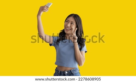 Cheerful young woman taking a selfie with a mobile phone and raising two fingers, Portrait of happy girl with bright face isolated on yellow background, Take a photo.