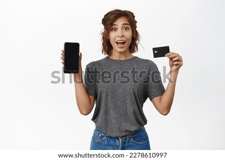 Portrait of girl posing with mobile phone and credit card, showing smartphone app screen, interface, standing over white background.