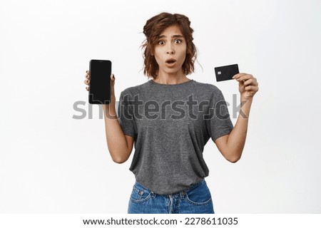 Worried girl showing her credit card, bank account on mobile phone screen, smartphone app, standing over white background.