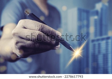 human hand holding a pen with a sparkling light at the nib. The background consists of blue skyscrapers slightly out of focus. The image suggests creativity and the power of writing.