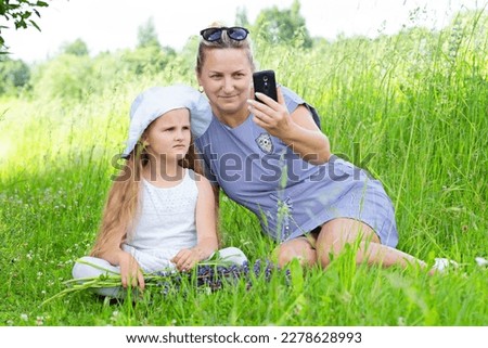 Attractive grandmother and cute granddaughter sit in the park and take a photo on the phone. Family selfies. Mom and daughter on the background of nature watching a mobile phone